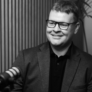 Dan Jennings, smiling and seated at a podcast-style microphone, wearing a suit jacket. Black and white portrait.