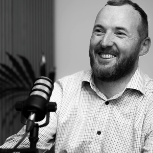 Mark Murphy, smiling and seated at a podcast-style microphone, wearing a checked shirt. Black and white portrait.